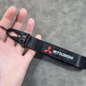 May include: Black fabric keychain with a black metal clasp and ring. The keychain features the Mitsubishi logo in red and the brand name in white. Designed for attaching keys or small items.