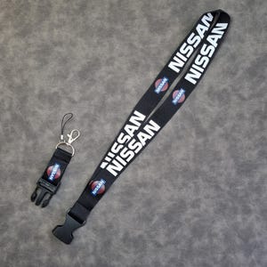 Puede incluir: Juego de cordón y llavero negros con el logotipo de Nissan en texto blanco y un emblema circular. El cordón tiene un cierre de plástico negro y el llavero tiene una hebilla de plástico negro y un cierre de metal.