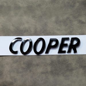 May include: Black and chrome "COOPER" emblem on a white strip. The letters are glossy and three-dimensional, with a reflective chrome finish on the "CO" letters. The emblem is likely for a car.