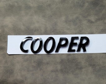 May include: Black and chrome "COOPER" emblem on a white strip. The letters are glossy and three-dimensional, with a reflective chrome finish on the "CO" letters. The emblem is likely for a car.
