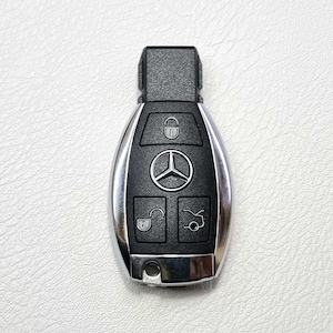 May include: A silver and black Mercedes-Benz car key. The key has a black top and a silver body with a black center section. The Mercedes-Benz logo is in the center, with lock and unlock buttons.