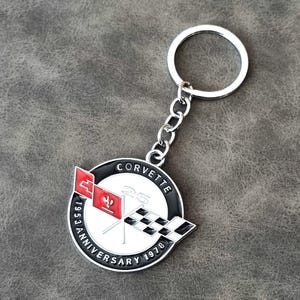 May include: Silver Corvette keyring with a round emblem. The emblem features the Corvette logo with red and black flags, the text "CORVETTE 25 ANNIVERSARY 1953-1978" in white and black, and a silver chain and key ring.