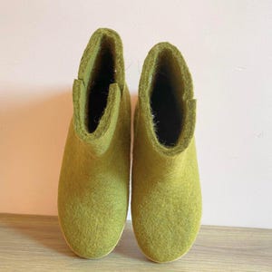 May include: A pair of olive green felt wool boots. The boots have a rounded toe and a short ankle height. The interior of the boots is dark. The boots are made of a soft, textured material and are set against a light pink background.