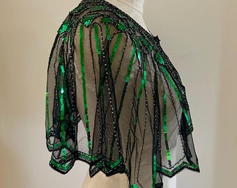 May include: Black sheer cape with vertical green sequin stripes. The cape is embellished with black beading along the neckline and hem. The garment is displayed on a white dress form.