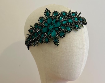 Puede incluir: Una diadema de cuentas de color verde azulado y negro con piedras grandes facetadas de color verde azulado dispuestas en un patrón floral. La diadema se muestra en una cabeza de maniquí.