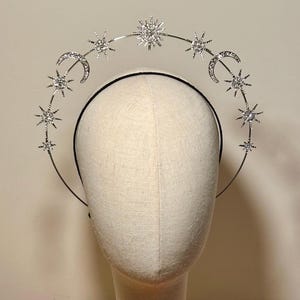 Könnte beinhalten: Ein silberner Himmels-Heiligenschein-Kranz mit Monden, Sternen und einem Sonnenstrahl-Design. Der Kranz wird auf einem Schaufensterpuppenkopf präsentiert. Der Heiligenschein ist aus Metall und mit glitzernden Verzierungen versehen. Der Kopf steht auf einem Holzständer.