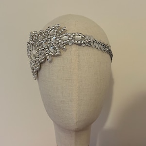 May include: A silver-toned headband with a floral design on the left side, adorned with clear rhinestones. The headband is displayed on a mannequin head with a neutral fabric covering.