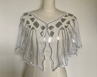 May include: A sheer, white cape-style top adorned with silver sequins. The cape features a V-neckline and a geometric pattern of silver sequins on the shoulders and vertical lines of sequins down the front.