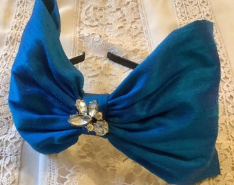 May include: A bright blue fabric headband featuring a large, oversized bow design. The center of the bow is embellished with a cluster of clear, faceted crystals that catch the light. The headband itself is a thin, dark, and flexible band.