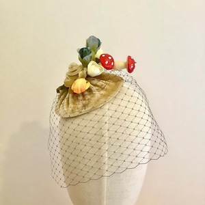Puede incluir: Un peque&ntilde;o sombrero de terciopelo beige con un adorno floral y de setas. El sombrero est&aacute; cubierto con un velo de tul negro.