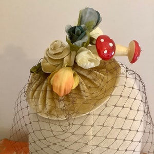Puede incluir: Un sombrero vintage con una base de terciopelo marr&oacute;n y un velo de red marr&oacute;n. El sombrero est&aacute; decorado con un grupo de flores de tela en tonos de blanco, amarillo, naranja, azul y verde. Dos adornos en forma de hongo rojo y blanco est&aacute;n unidos a las flores.