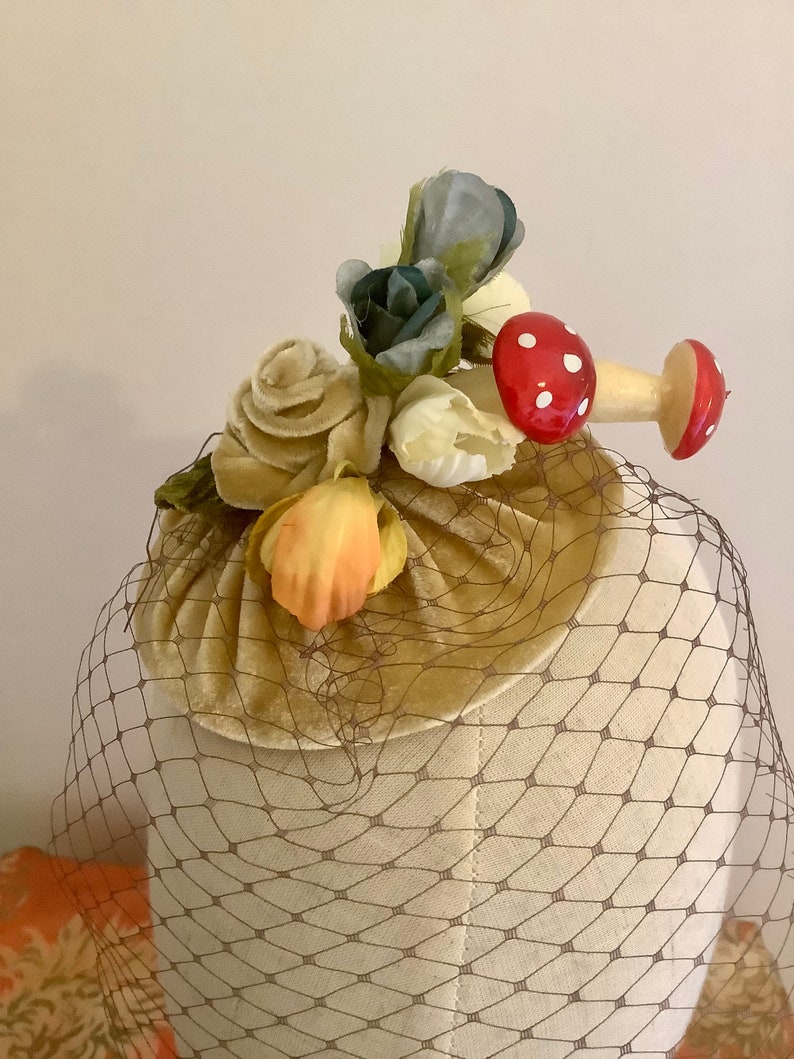 Puede incluir: Un sombrero vintage con una base de terciopelo marr&oacute;n y un velo de red marr&oacute;n. El sombrero est&aacute; decorado con un grupo de flores de tela en tonos de blanco, amarillo, naranja, azul y verde. Dos adornos en forma de hongo rojo y blanco est&aacute;n unidos a las flores.