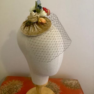 Puede incluir: Un peque&ntilde;o sombrero de terciopelo beige con un adorno floral y de setas. El sombrero est&aacute; adornado con flores blancas, amarillas y azules, as&iacute; como una seta roja. El sombrero est&aacute; en una cabeza de maniqu&iacute; blanca.