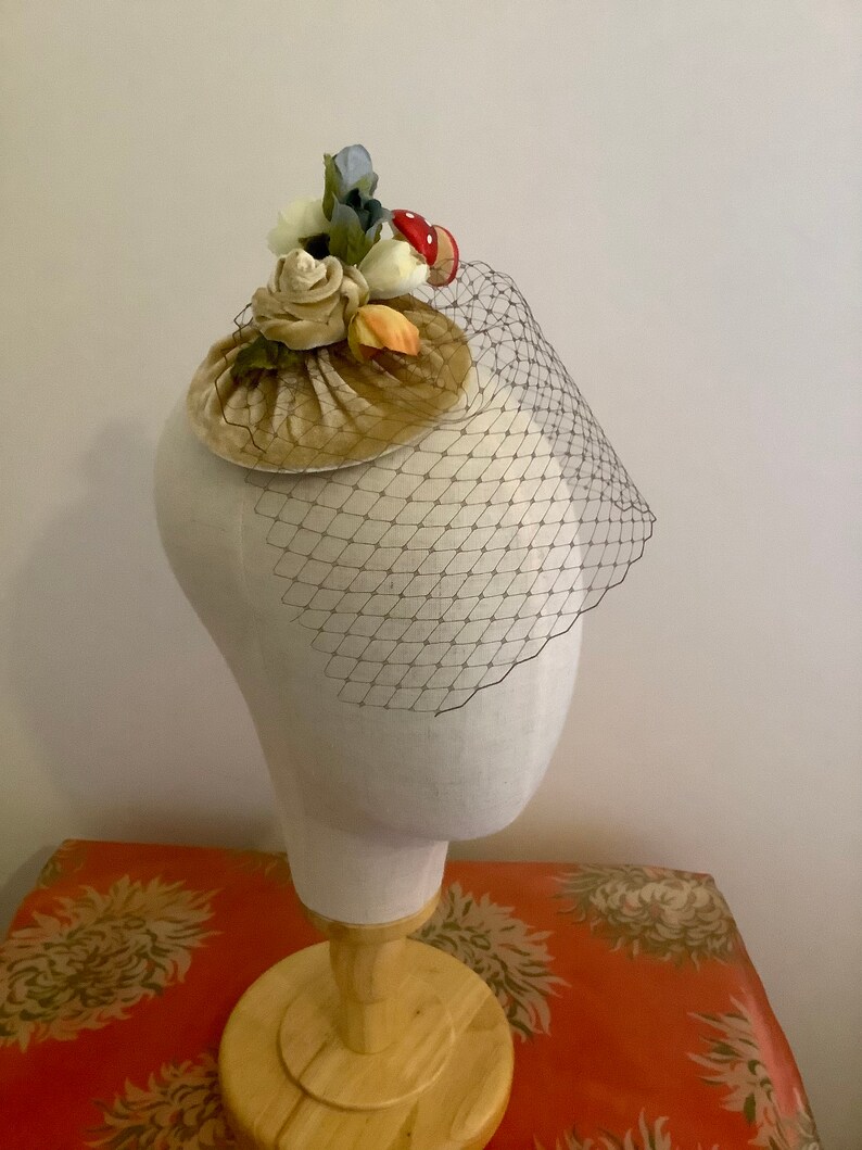 Puede incluir: Un peque&ntilde;o sombrero de terciopelo beige con un adorno floral y de setas. El sombrero est&aacute; adornado con flores blancas, amarillas y azules, as&iacute; como una seta roja. El sombrero est&aacute; en una cabeza de maniqu&iacute; blanca.