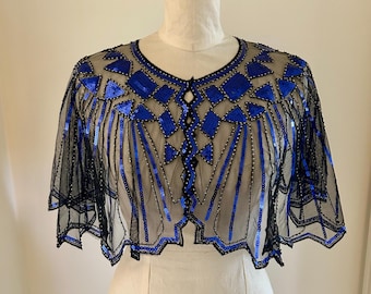 May include: Black sheer capelet with blue sequin and bead embellishments. The capelet features a geometric design with a scalloped hemline. The garment is displayed on a mannequin.