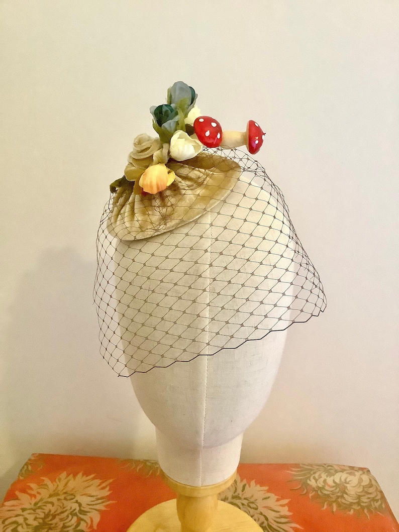 Puede incluir: Un peque&ntilde;o sombrero de terciopelo beige con un adorno floral y de setas. El sombrero est&aacute; cubierto con un velo de tul negro.