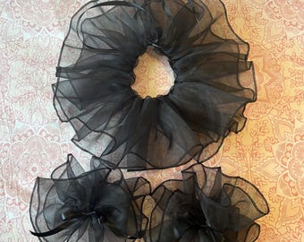 May include: A set of three black hair accessories made of sheer, ruffled organza fabric. The collection includes one large, circular scrunchie and two smaller, matching hair ties, each featuring delicate ribbon accents and layered, voluminous folds.