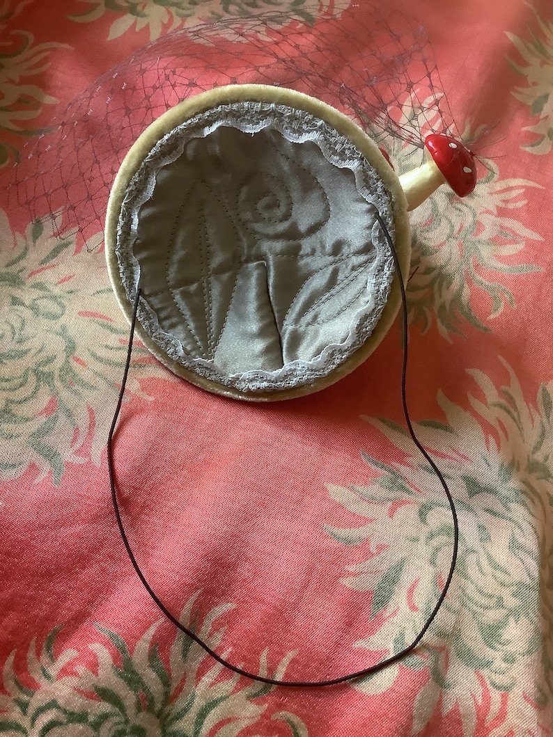 Puede incluir: Un sombrero de terciopelo blanco con un dise&ntilde;o de hongo rojo y blanco. El sombrero tiene una banda el&aacute;stica negra y un velo de red blanca.
