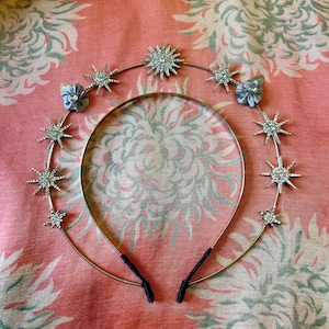Könnte beinhalten: Ein silbernes Stirnband mit Stern- und Blumendetails. Das himmlische Kopfstück hat mehrere sternförmige Verzierungen mit klaren Kristallakzenten. Zwei kleine Stoffblumen sind ebenfalls angebracht. Das Stirnband wird auf einem rosa geblümten Hintergrund präsentiert.