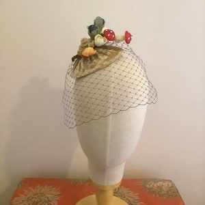 Puede incluir: Un peque&ntilde;o sombrero de terciopelo marr&oacute;n con velo. El sombrero est&aacute; decorado con conchas marinas, flores y setas rojas.