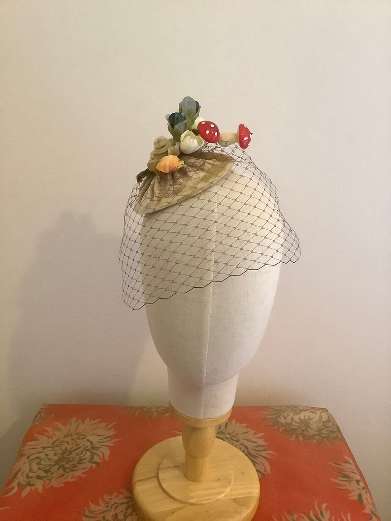 Puede incluir: Un peque&ntilde;o sombrero de terciopelo marr&oacute;n con velo. El sombrero est&aacute; decorado con conchas marinas, flores y setas rojas.