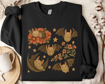 May include: Black sweatshirt with a playful design of bats, flowers, and stars. The word "Black" is written in cursive. The bats are brown with outstretched wings, and the flowers are orange. A charming and cosy design.