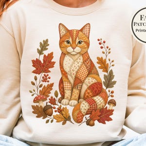 Puede incluir: Una sudadera beige de cuello redondo con un gráfico de un gato con patrón de falso patchwork en tonos naranja, marrón y crema. El gato está rodeado de hojas de otoño, bellotas, bayas y setas. Una insignia circular dice FAUX PATCHWORK Printed Design.
