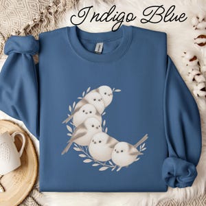 May include: An indigo blue crewneck sweatshirt featuring a graphic print of five small, round, light-coloured birds perched together in a crescent moon shape surrounded by delicate white leafy branches. The text Indigo Blue is printed above.