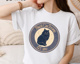 May include: White t-shirt featuring a circular graphic for the "Black Cat Society." The design includes a black cat silhouette, a crescent moon, and the text "Est. 1692." The word "White" is written in cursive at the top left.
