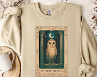 May include: Sand-colored sweatshirt featuring a tarot card design. The card depicts an owl with a crescent moon and stars, framed by ornate columns. The text "THE HIGH PRIESTESS" is at the bottom. The sweatshirt has a crew neck and long sleeves.