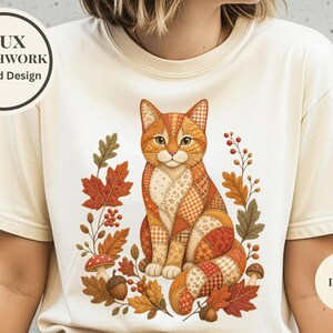 Könnte beinhalten: Ein elfenbeinfarbenes T-Shirt mit einem aufgedruckten Faux-Patchwork-Design einer orangefarbenen Katze, umgeben von Herbstblättern, Beeren, Pilzen und Eicheln. Die Grafik zeigt verschiedene Muster in warmen Tönen. Der Text lautet FAUX PATCHWORK Printed Design und IVORY.