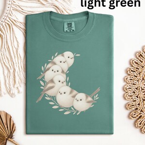 Könnte beinhalten: Hellgrünes T-Shirt mit einem Motiv einer Gruppe weißer Schwanzmeisen, die in Form eines Halbmondes angeordnet sind. Das T-Shirt hat ein Comfort Colors-Etikett.