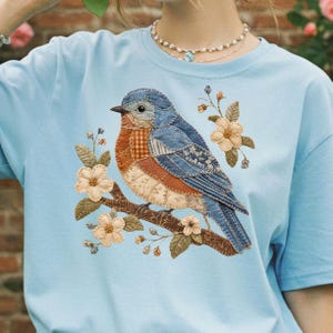 May include: Pale blue t-shirt with a patchwork design of a bluebird on a branch with flowers. The bird is made from denim fabrics, and the flowers are beige. The shirt is made of a soft, comfortable material.