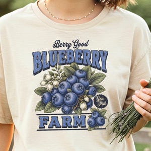 May include: A cream-colored t-shirt featuring a graphic print of blueberries with green leaves and white flowers. The text reads Berry Good BLUEBERRY FARM You Pick in blue and black lettering. The design has a vintage, distressed aesthetic.