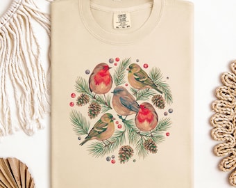 May include: Ivory-colored t-shirt featuring a circular design of six birds perched on pine branches with pinecones and berries. The shirt has a crew neck and the words "COMFORT COLORS" are printed on the label.