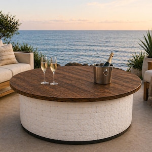 May include: A round outdoor coffee table with a dark wood top and a white brick-style base. Two champagne flutes and a silver ice bucket with a champagne bottle are on the table. The table is surrounded by outdoor furniture and the ocean.