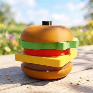 Puede incluir: Un juguete de madera en forma de hamburguesa con capas marrones, amarillas, rojas y verdes. La parte superior del pan tiene una pequeña clavija negra. El juguete está sobre una superficie de madera.