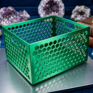 May include: A vibrant green metal storage crate featuring a honeycomb design. The crate is rectangular with side handles. It is displayed on a silver surface. The crate's dimensions are approximately 25 cm x 25 cm x 15 cm.