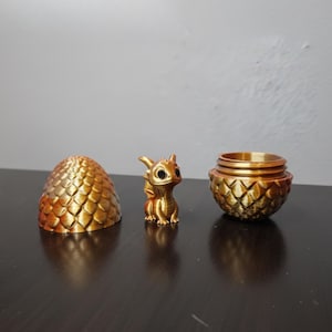 Puede incluir: Una figura de dragón dorado con ojos grandes se encuentra junto a una cáscara de huevo y un recipiente a juego. La cáscara de huevo y el recipiente tienen una textura similar a las escamas. El dragón tiene pequeñas alas y está sentado.
