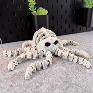 May include: A white, articulated octopus skeleton figurine. The octopus has a skull-like head with large eye sockets and segmented tentacles. The figurine is displayed on a grey surface.