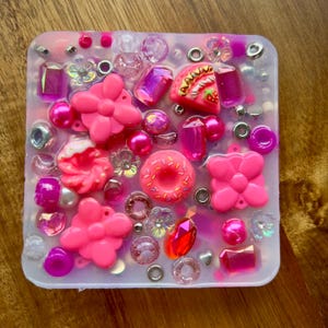 Könnte beinhalten: Ein quadratisches, klares Harztablett gefüllt mit rosa, roten und weißen Charms. Die Charms umfassen Blumen, Donuts, eine Erdbeerscheibe und verschiedene andere Formen.