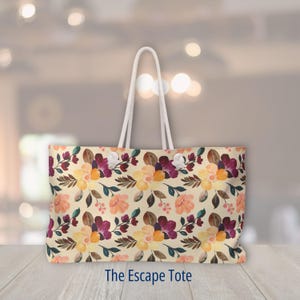 May include: A rectangular tote bag with a floral pattern in shades of yellow, purple, and orange on a cream background. The bag has white rope handles and the text "The Escape Tote" at the bottom.