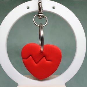 Op de afbeelding: Een rode hartvormige sleutelhanger met een gebroken hartontwerp en een zilveren sleutelring. De sleutelhanger hangt aan een witte ronde standaard.