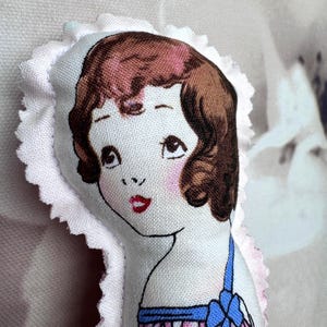 May include: A handmade fabric doll with a vintage design. The doll features a printed face with brown hair, rosy cheeks, and red lips. It is wearing a blue and pink dress and is surrounded by a white, ruffled trim.