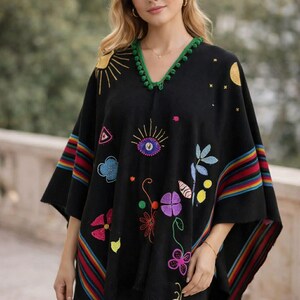 May include: A black poncho featuring a green pom-pom trim along the V-neckline. The garment is decorated with colorful embroidered motifs including a sun, moon, stars, an eye, and various flowers. The sleeves and hem have multi-colored stripes.