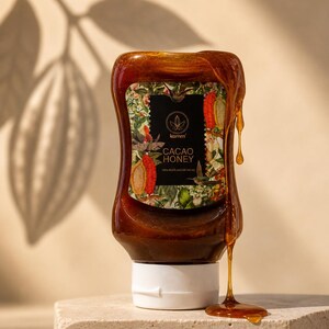 以下が含まれることがあります： フローラルなラベルと「Cacao Honey」の文字が入ったカカオハニーのボトル。蜂蜜は濃い茶色で、ボトルの側面を伝って滴り落ちています。ボトルは石の表面に置かれています。