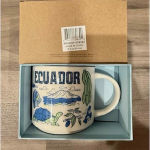 Könnte beinhalten: Eine Keramiktasse mit dem Wort "ECUADOR" in blauer Schrift, mit Illustrationen der lokalen Flora und Fauna. Die Tasse wird in einer hellblauen Box mit einem braunen Kartondeckel präsentiert. Fassungsvermögen: 414 ml.