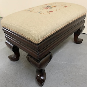 May include: A wooden footstool with a floral embroidered top and four carved legs. The footstool is upholstered in a beige fabric.
