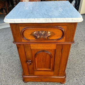 May include: A wooden nightstand with a marble top. The nightstand has a drawer with ornate carvings and a cabinet door. The wood has a warm, reddish-brown tone. The marble top is light gray with white veining.
