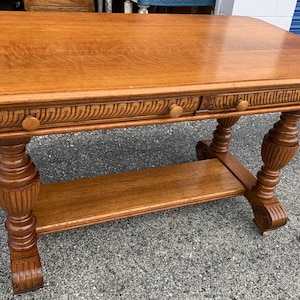 May include: A vintage wooden table with a carved design and two drawers. The table has a shelf and turned legs.